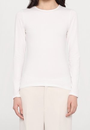 Mujer con una camiseta blanca lisa y ajustada de manga larga y pantalones beige claro, de pie frente a un fondo blanco.