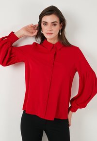 Red blouse featuring long, puffed sleeves, a button-front closure, and a collar. The fabric appears smooth with a subtle sheen.