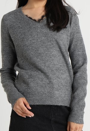 Femme portant un pull gris doux à col en V avec bordure en dentelle et pantalon noir, debout devant un fond clair uni.