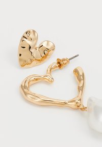 Pendientes en tono dorado con forma de corazón y una textura ondulada, acentuados por un detalle de perla blanca lisa.