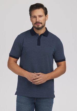 Mężczyzna w granatowej koszulce polo z krótkim rękawem, z ciemnym kołnierzykiem i mankietami, stoi ze złączonymi rękami na neutralnym tle.