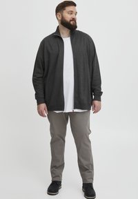 Giacca zip grigia con colletto alto, indossata sopra una maglietta bianca. Pantaloni beige e scarpe nere completano l'outfit. Il tessuto appare morbido.