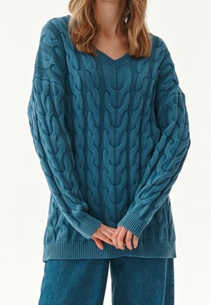 Frau trägt einen türkisblauen, grob gestrickten V-Ausschnitt-Pullover mit Zopfstrickmuster und dazu passende blaue Hosen mit weitem Bein, die Hände locker vor dem Körper verschränkt.