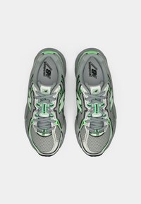 Pelēkas sporta kurpes ar zaļām detaļām, acs materiāla augšdaļa, atstarojošas detaļas un amortizētas zolītes. Redzama šņorēšanas sistēma un logo mēlītē.