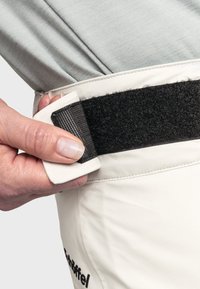 Weiße Cargo-Hosen verfügen über einen schwarzen Klettverschluss. Eine Hand greift den Verschluss und zeigt die Materialien sowie das funktionale Design.