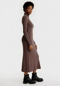 Langes, geripptes braunes Kleid mit hohem Kragen, figurbetonter Silhouette und ausgestellt geschnittenem Saum, kombiniert mit schwarzen klobigen Stiefeletten.