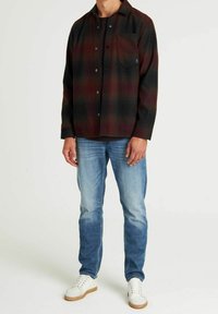 Camicia da uomo in flanella a quadri rosso scuro e nero, abbottonata con una tasca sul petto. Abbinata a jeans azzurri chiaro e sneakers bianche da ginnastica.