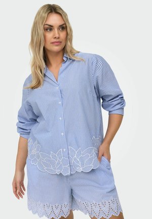 Naine, kellel on seljas sinise ja valge triibuline nööbitav särk ning sobivad lühikesed püksid valgete lillelistest pitsidest detailidega, seisab ühe käega taskus.