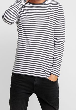 Homme portant une chemise à manches longues rayée horizontalement noir et blanc avec un petit logo sur la poitrine et un pantalon noir, main dans la poche et touchant son cou.