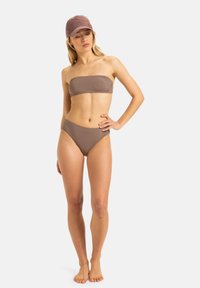 Brauner Bandeau-Bikinitop kombiniert mit hochgeschnittenen Bikinihöschen aus strukturiertem Stoff. Getragen mit einer passenden Mütze und barfuß.