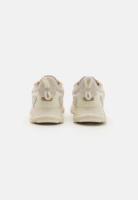 Beige sneakers with a textured fabric upper, smooth leather accents, and a cushioned sole. Features a pull tab on the heel for easy wear.