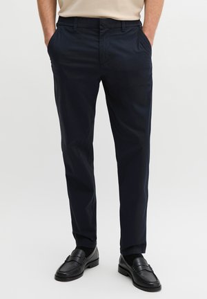 Mężczyzna ubrany w granatowe, dopasowane spodnie i czarne skórzane mokasyny, z rękami w kieszeniach na jednolitym tle.