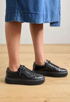 Jambes portant des baskets compensées noires avec des fermetures éclair latérales et des lacets, debout sur un sol en bois, associées à une jupe en denim bleu.