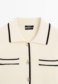 Pullover in cotone con base color crema, rifiniture nere e colletto polo. Presenta tre bottoni rotondi e due piccole tasche con accenti neri.