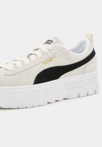 Baskets blanches Puma avec bande latérale noire, semelle épaisse, lacets blancs et logo Puma doré sur tige en daim.