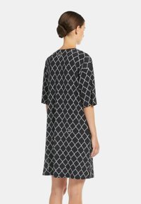 Donna con capelli castani raccolti in un basso chignon, che indossa un vestito nero al ginocchio con motivi geometrici bianchi, in piedi di spalle contro uno sfondo semplice.