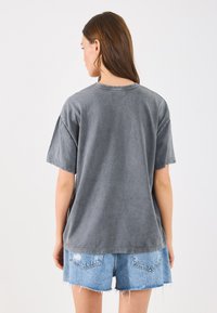 Graues, übergroßes Baumwoll-T-Shirt mit kurzen Ärmeln, das einen runden Ausschnitt und eine weiche, leicht strukturierte Oberfläche aufweist. Kombiniert mit Jeansshorts.
