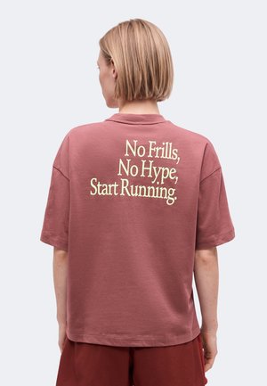 Persona con capelli biondi corti che indossa una maglietta larga bordeaux con la scritta "No Frills, No Hype, Start Running." sul retro.