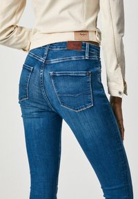 Blue denim jeans with a slim fit, featuring back pockets and a brown leather label on the waistband. The fabric has a slight fade and texture.
