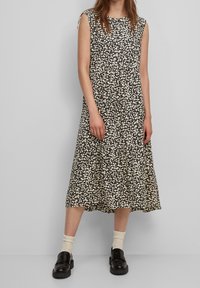 Vestido con estampado floral en negro y crema, sin mangas, con un dobladillo con volantes, hasta la rodilla, hecho de una tela ligera. Combinado con zapatos negros gruesos.
