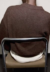 Smeši smeđi pleteni pulover s okruglim izrezom, nosi se preko bijelih hlača, sjedi na stolici od metalnog okvira nasuprot jednostavne pozadine.