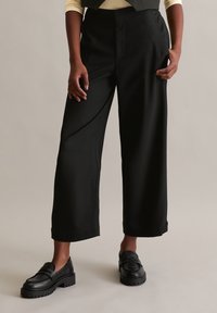 Schwarze weit geschnittene Hosen aus glattem Stoff, mit tailliertem Bund, seitlichen Taschen und geradem Saum. Kombiniert mit schwarzen Slippern.