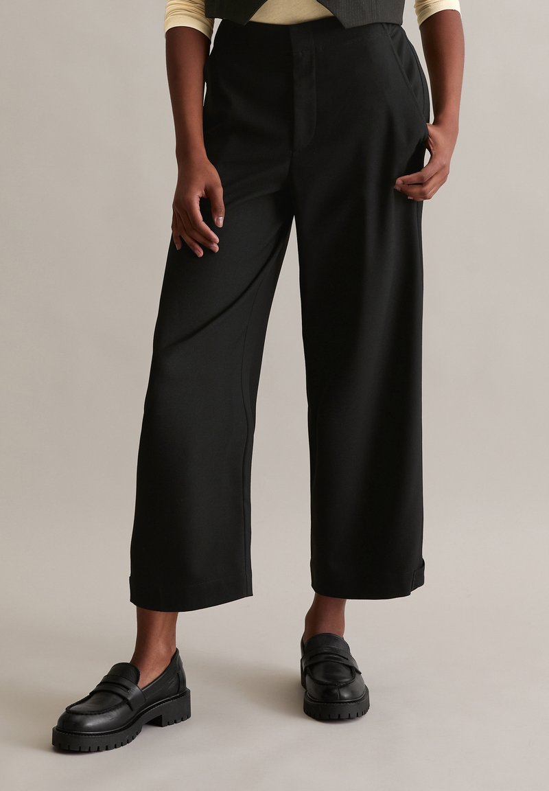 Schwarze weit geschnittene Hosen aus glattem Stoff, mit tailliertem Bund, seitlichen Taschen und geradem Saum. Kombiniert mit schwarzen Slippern.