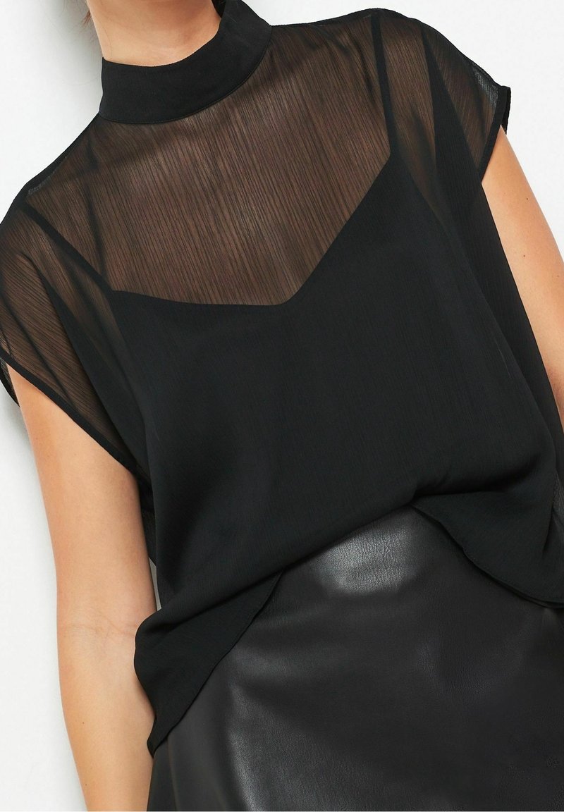 Femme portant un chemisier noir transparent à col montant superposé sur un caraco noir, associé à une jupe en cuir noire.
