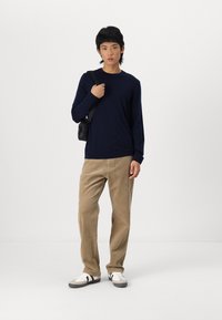 Jūrininkų ilgos rankovės marškiniai, smėlio spalvos velveto kelnės, juoda mažoji petneša ir baltos sportinės bateliai su juodais akcentais. Paprasta fono.