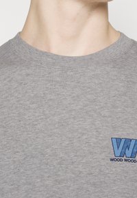 Grå bomullströja med rund halsringning, prydd med en blå broderad logotyp på vänster sida, mjuk textur och enkel design.