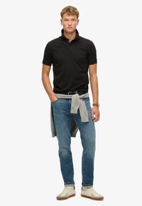 Polo nero con colletto, maglione grigio chiaro annodato in vita, jeans blu e sneakers crema con suole marroni.