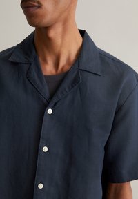 Korte mouw, donkerblauwe shirt van lichte stof, met een gelapte kraag, vijf witte knopen en een relaxte pasvorm. Twillstructuur zichtbaar.
