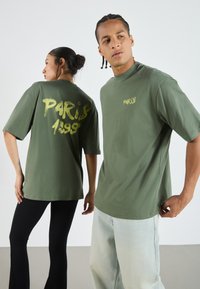 Oliven grønne oversized t-shirts med "PARIS 1998" trykt i gul på ryggen og foran, parret med sorte bukser og lyseblå jeans.