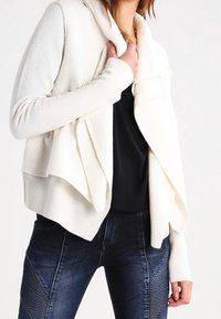 Mjuk krämfärgad kofta med öppen front, som har ett strukturerat mönster på den övre halvan och ribbade ärmar. Matchad med mörk denimm jeans.