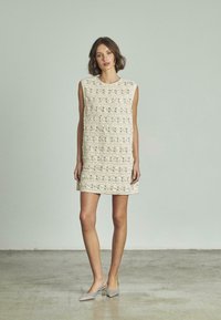 Spitzenkleid in creme mit rundem Ausschnitt und ärmellosem Design, das strukturierte Muster aufweist. Kombiniert mit silbernen Absätzen.
