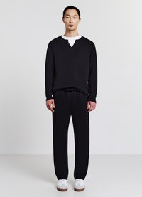Pullover nero a maniche lunghe con scollo a V e dettagli di bottoni, abbinato a pantaloni neri con coulisse e sneakers bianche, su uno sfondo neutro.