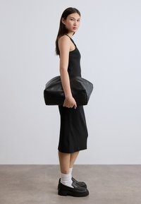 Μαύρη δερμάτινη τσάντα με μαλακό, στρογγυλεμένο σχήμα, κρατημένη στο χέρι. Το μοντέλο φοράει εφαρμοστό μαύρο φόρεμα και χοντρά μαύρα παπούτσια με λευκές κάλτσες.