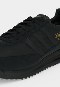 Μαύρο αθλητικό παπούτσι Adidas SL72 με σουέτ λεπτομέρειες, ρίγες με ζιγκ-ζαγκ άκρες, μαύρα κορδόνια και χρυσό λογότυπο στο πλάι, σε λευκό φόντο.