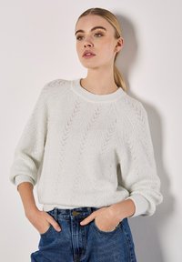 Witte gebreide trui met een gestructureerd visgraatpatroon, ronde halslijn en verlaagde schouders, gecombineerd met donkerblauwe high-waisted jeans.