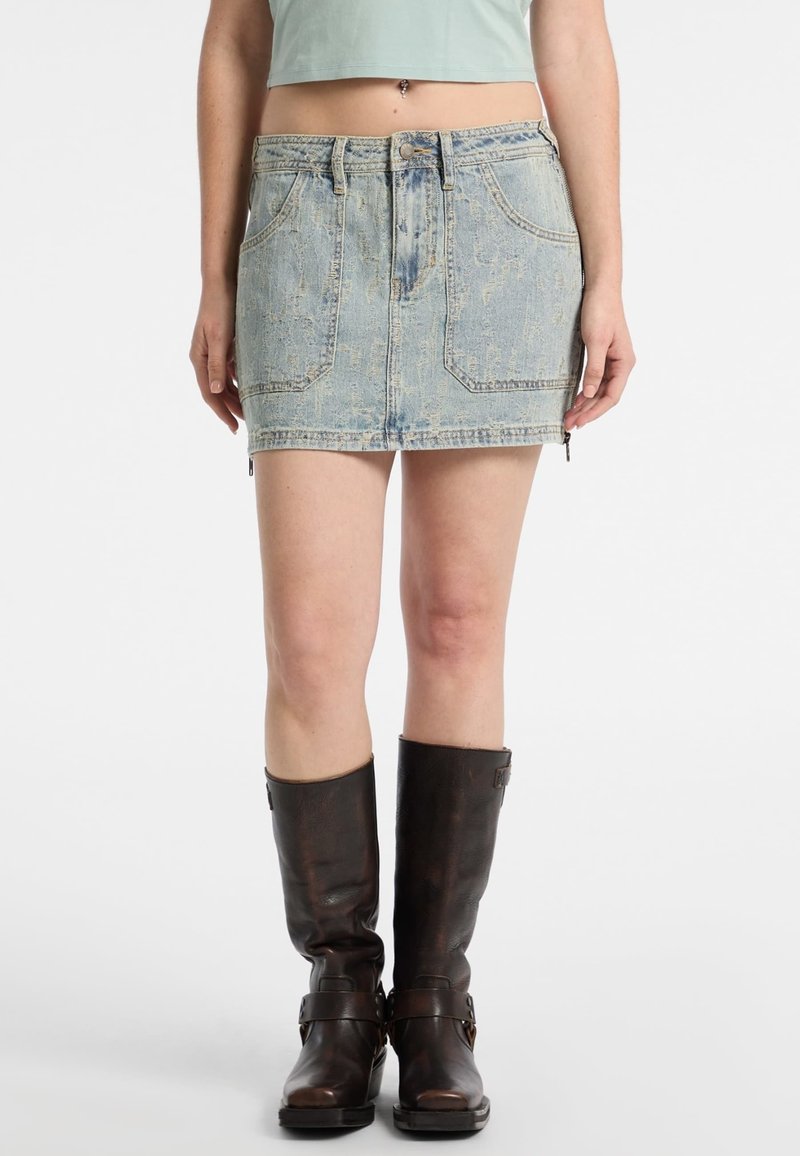 Denimowa mini spódnica o wyblakłym niebieskim odcieniu, z przednimi kieszeniami i zamkiem błyskawicznym. Nosi się ją z długimi brązowymi skórzanymi botkami z akcentami klamer.