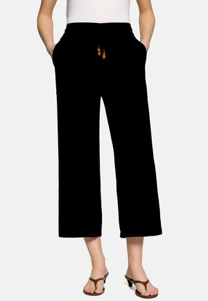 Femme portant un pantalon large noir coupé avec un cordon perlé, un haut blanc et des sandales marron à talons, mains dans les poches, debout.