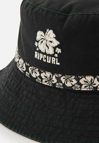 Chapeau seau noir avec une finition texturée, arborant un design floral blanc brodé et le logo "RIPCURL", complété par un bandeau à motifs.