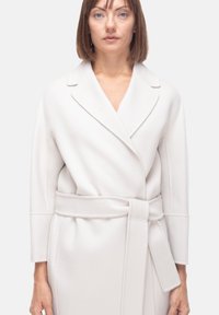 Cappotto avvolgente grigio chiaro realizzato in tessuto morbido, con vita in vita, colletto a scialle e maniche di lunghezza medio-corta con dettagli di cuciture minime.