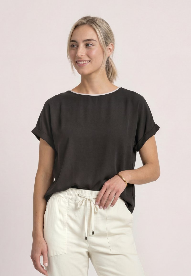 Jeune femme aux cheveux blonds attachés en queue de cheval basse, portant un t-shirt noir ample à manches courtes et un pantalon blanc à cordon, souriante et regardant vers la droite.