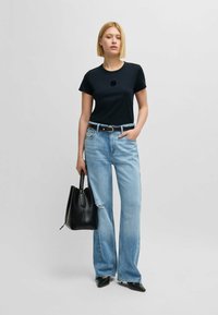 Czarny t-shirt z krótkim rękawem z teksturalnym detalem w kształcie kwiatu, zestawiony z jasnoniebieskimi spodniami typu wide-leg i czarną torbą typu bucket. Czarne buty na szpilkach.