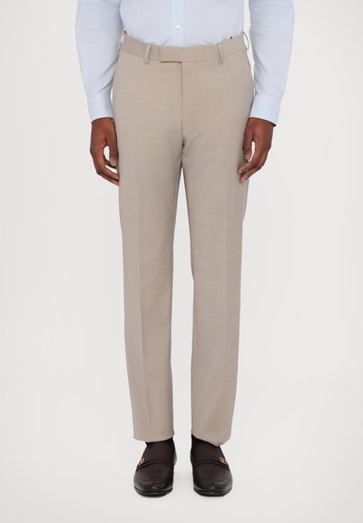 Pantaloni beige su misura con una texture liscia, davanti piatto e tasche laterali. Indossati con una camicia azzurro chiaro e mocassini scuri.