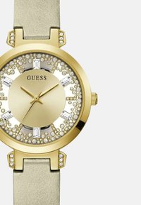 Orologio dorato con quadrante rotondo, dettagli in cristallo e cinturino in pelle beige. Presenta un quadrante grande con una finitura lucida e lancette metalliche.