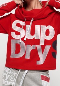 Sudadera con capucha roja con gran texto perforado "Sup" y "Dry" en blanco y gris. Tiene cordones y un corte holgado.
