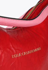 Živá červená kabelka má růžovou rukojeť a zlaté logo "LOLA CASADEMUNT", což podtrhuje její stylový a moderní design.