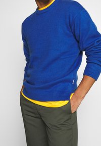 Niebieski sweter z dzianiny z okrągłym dekoltem, z prążkowanymi mankietami i dołem, nałożony na żółtą koszulkę, w zestawieniu z oliwkowymi spodniami.